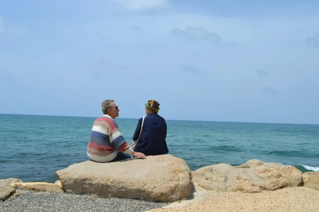 Couple de seniors assis sur des rochers face à la mer, partageant un moment calme.